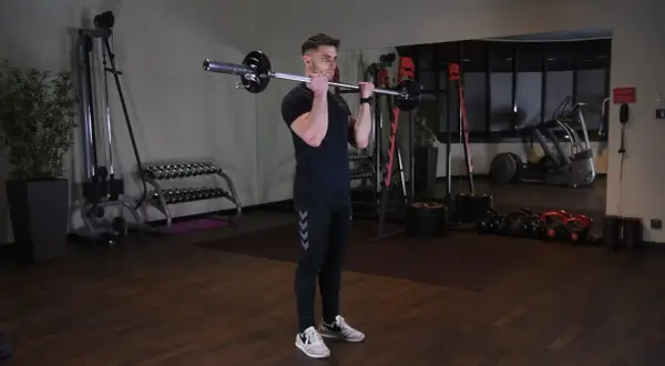Mężczyzna wykonuje ćwiczenie barbell curl w siłowni. W tle widać sprzęt do ćwiczeń.