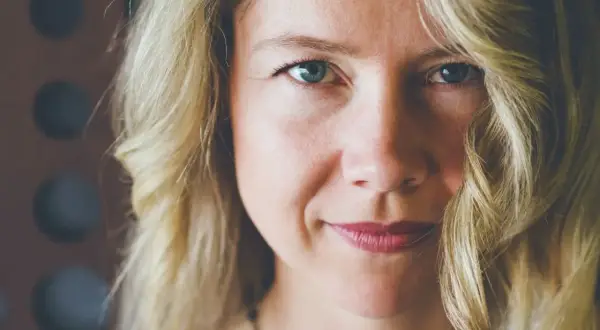 Zbliżenie na twarz kobiety z blond włosami. Delikatny uśmiech, lekko opadające policzki, niebieskie oczy.