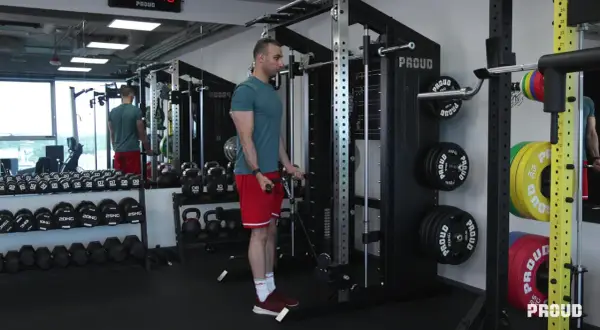 Mężczyzna wykonuje ćwiczenia na biceps przy użyciu wyciągu. W tle widoczne są sztangi, hantle i talerze obciążeniowe.