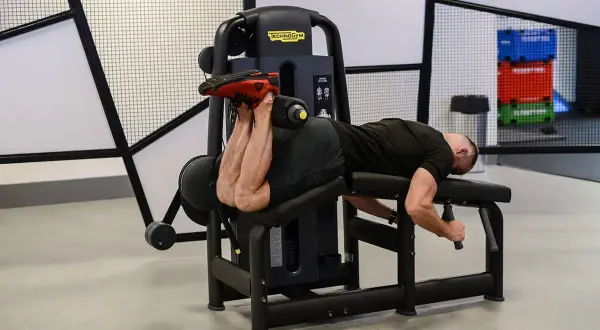 Mężczyzna wykonuje ćwiczenie na maszynie do uginania nóg leżąc (hamstring curl), budując siłę tylnej części uda.