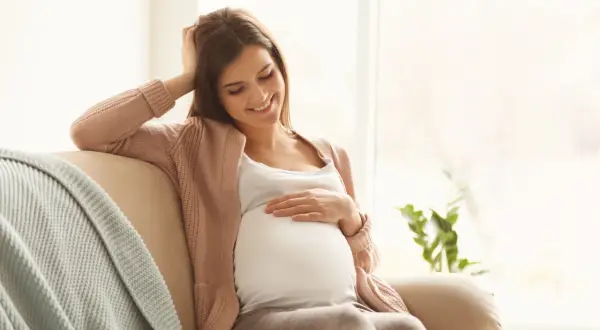 Uśmiechnięta ciężarna kobieta siedzi na kanapie, delikatnie dotykając brzucha. Myśli o bezpiecznym oczyszczaniu wodorowym w ciąży.