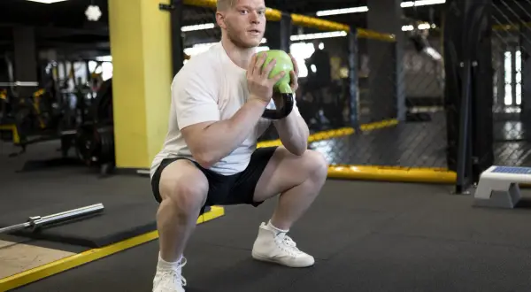 Mężczyzna wykonuje goblet squats z zielonym kettlebellem w siłowni.