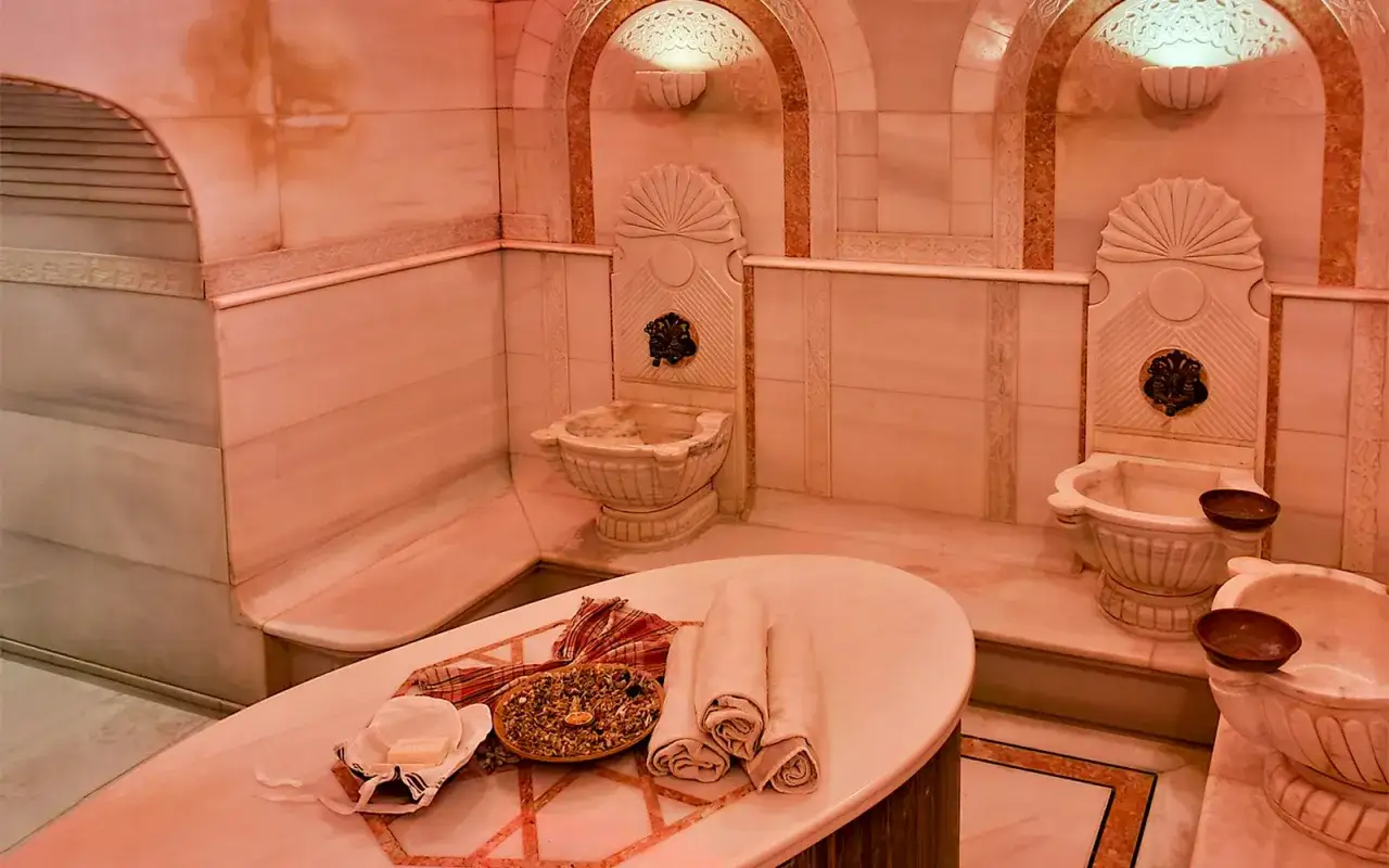 Luksusowy hammam z marmurowymi umywalkami i ozdobnymi detalami. Na stole leżą ręczniki i mydło, gotowe na relaksującą kąpiel.